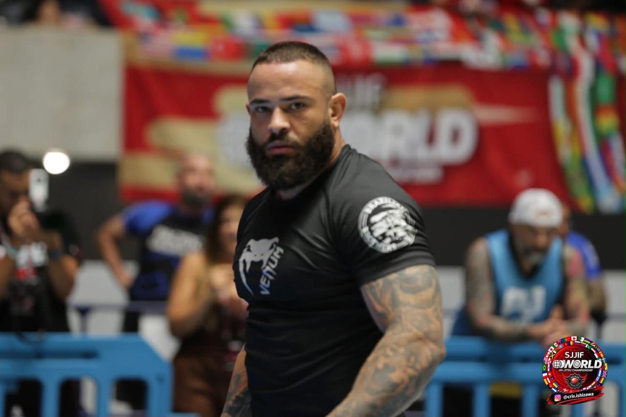 Uriel Meneses de rashguard preta encarando a câmera na área de luta do Mundial SJJIF 2026, em Tóquio, com o painel do campeonato e bandeiras ao fundo