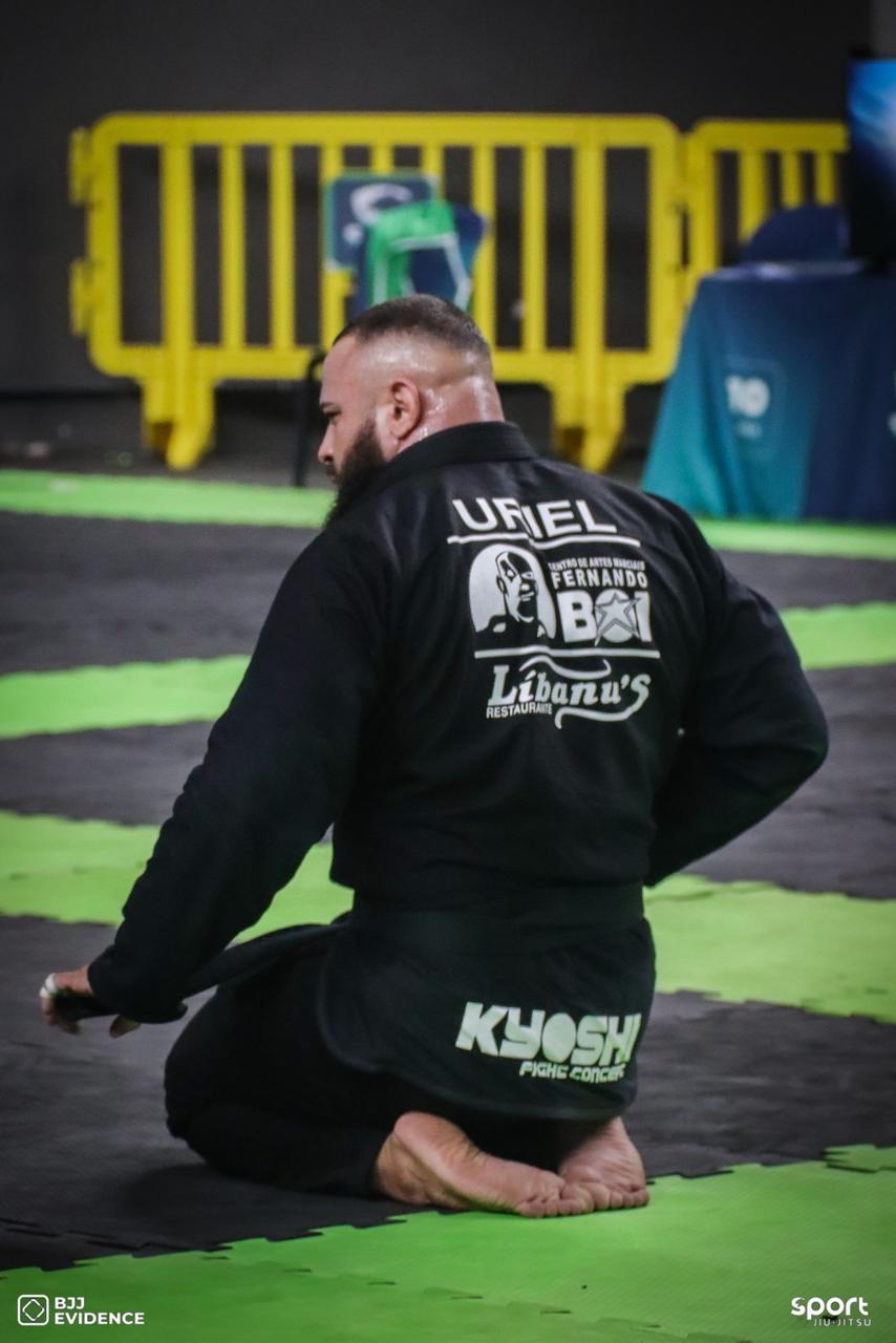 Uriel Meneses ajoelhado no tatame, de costas, com Fernando Boi, Libanus e Kyoshi bordados no kimono preto de competição